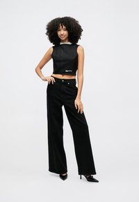 Čierny crop top z denimu so štvorcovým výstrihom a detailom loga, doplnený širokými čiernymi nohavicami z denimu. Modelka má na nohách čierne lodičky s ostrou špičkou.