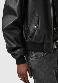 Czarna skórzana kurtka typu bomber z prążkowanym kołnierzem i mankietami, wyposażona w boczną kieszeń, zestawiona z szarymi dżinsami. Zbliżenie na detale.