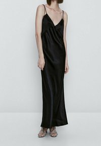 Robe maxi en satin noir avec un décolleté plongeant, de fines bretelles ajustables et une silhouette fluide. Texture lisse avec des détails de design minimalistes.