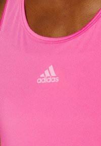Vaaleanpunainen urheilullinen toppi, joka on valmistettu sileästä kankaasta. Siinä on pyöreä kaula-aukko ja edessä on valkoinen Adidas-logo.