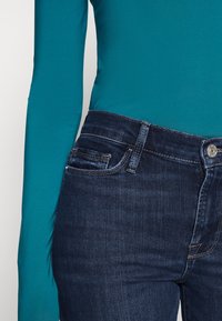 Pantalones vaqueros de mezclilla azul con un lavado medio, que presentan un cierre de cremallera, cinco bolsillos y costuras de contraste. Combinados con una blusa de manga larga en color teal.