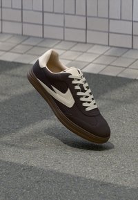 Dunkelbrauner und beiger Sneaker mit weißen Schnürsenkeln schwebt über strukturiertem Pflaster neben einer gefliesten Wand.