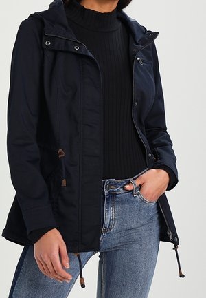Woman wearing a navy hooded jacket over a black ribbed turtleneck sweater and blue jeans, with one hand in pocket.