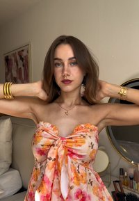 Jeune femme avec des cheveux bruns à épaule, portant une robe sans bretelles à motifs floraux, des bijoux en or, ajustant ses cheveux dans une chambre avec du maquillage sur une coiffeuse.