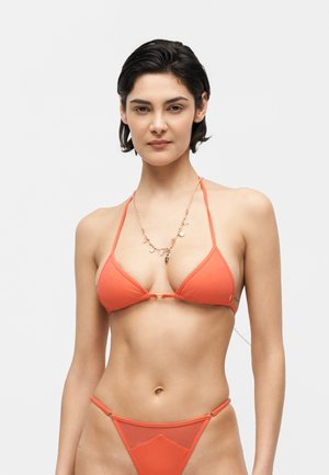 Femeie cu păr scurt și întunecat, purtând un top de bikini portocaliu triunghiular și chiloți asortați, pozând pe un fundal alb simplu.