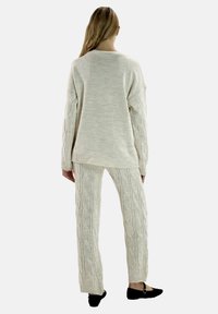 Kremfarvet strikket sweater med sænkede skuldre, kombineret med matchende kabelstrikkede bukser. Begge har teksturerede mønstre og ribstrikkede manchetter.