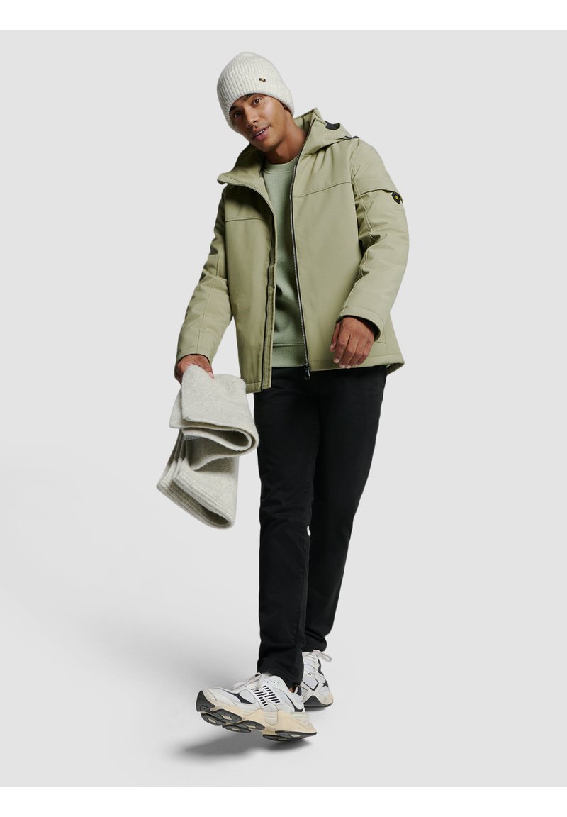 Veste matelassée vert clair avec capuche, pull gris, pantalon noir et baskets blanches. Tient deux couvertures gris clair. Bonnet tricoté blanc.