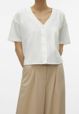 Persoon die een wit kort mouw overhemd draagt met knopen en een V-hals, en beige plissé wijde broek met hoge taille.
