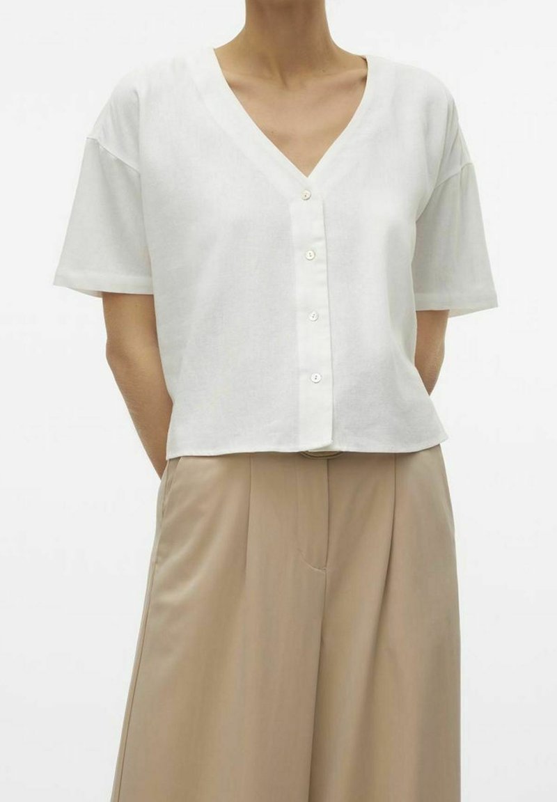 Personne portant une chemise blanche à manches courtes avec boutons et col en V, ainsi qu’un pantalon beige taille haute plissé à jambes larges.