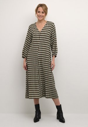 Maxikleid mit V-Ausschnitt, langen Ärmeln und einem geometrischen schwarz-beigen Muster. Kombiniert mit schwarzen, knöchelhohen High Heels.