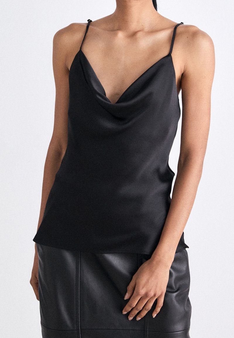 Femme portant un haut sans manches en satin noir avec de fines bretelles et un décolleté drapé, assorti à une jupe en cuir noire.