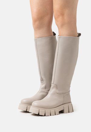 Knee-high beige leather boots with a smooth texture, rounded toe, and chunky rubber sole featuring horizontal ridges. Minimalist design detail.