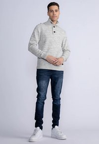 Hellgrauer Strickpullover mit hohem Kragen, zwei Knöpfen und gerippter Textur, kombiniert mit dunkelblauen Jeans und weißen Sneakers.