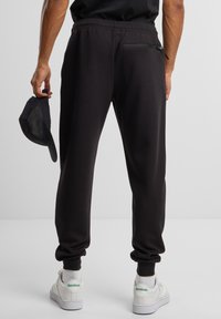 Joggers negros con cintura elástica, puños en los tobillos y un bolsillo con cremallera en la parte trasera. Combinados con zapatillas blancas con detalles en verde.
