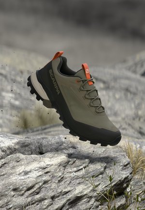 Olivgrüner und schwarzer Wanderschuh mit orangefarbenen Details, der über felsigem Gelände im Freien schwebt.
