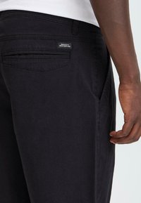 Svarta denimbyxor på en person med en sidoficka och en bakficka synlig, med en liten etikett som läser "DEDIKERADE TILL DENIMPERFEKTION."