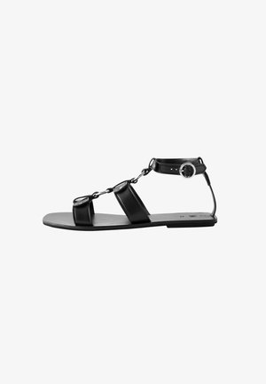 Sandalia de cuero negro con tres tiras y un detalle de cadena, que presenta una correa para el tobillo ajustable con un hebilla circular y suela plana.
