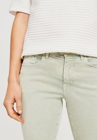 Vaqueros verde claro con una textura suave, con un diseño de cinco bolsillos y cierre frontal de botones, combinados con una camiseta blanca acanalada.