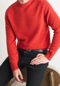 Roter Strickpullover mit Rundhalsausschnitt und gerippten Bündchen, kombiniert mit schwarzen Hosen. Die Textur wirkt weich und warm, geeignet für lässige Kleidung.