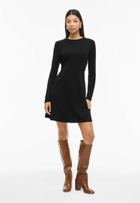 Robe en tricot côtelé noire avec manches longues, couture à la taille et jupe évasée. Portée avec des bottes marron montant jusqu'aux genoux, à talons empilés.