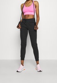 Schwarze Sweatpants mit elastischen Bündchen, kombiniert mit einem pinkfarbenen Sport-BH mit weißem Logo. Die Person hält ein Springseil. Pinkfarbene Sportschuhe.
