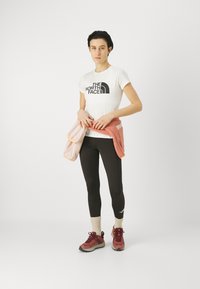 Biała koszulka z krótkim rękawem z czarnym logo, czarne legginsy i czerwone buty trekkingowe. Różowy sweter związany w talii. Neutralne tło.