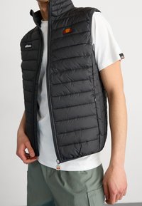 Lekka czarna kamizelka pikowana z pionowymi przeszyciami, wysokim kołnierzem i przednim zamkiem błyskawicznym. Posiada małe logo na piersi i przy zatrzasku zamka.