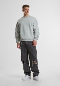 Hellgrauer Sweatshirt mit Rundhalsausschnitt, kombiniert mit schwarzen, ausgewaschenen, weiten Jeans mit großen Rissen. Weiße Turnschuhe, lässiger Look.