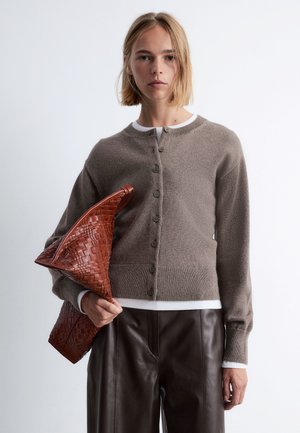 Brown knit cardigan with round neckline, five buttons, and ribbed cuffs. Woven red handbag held in right hand, leather trousers visible.