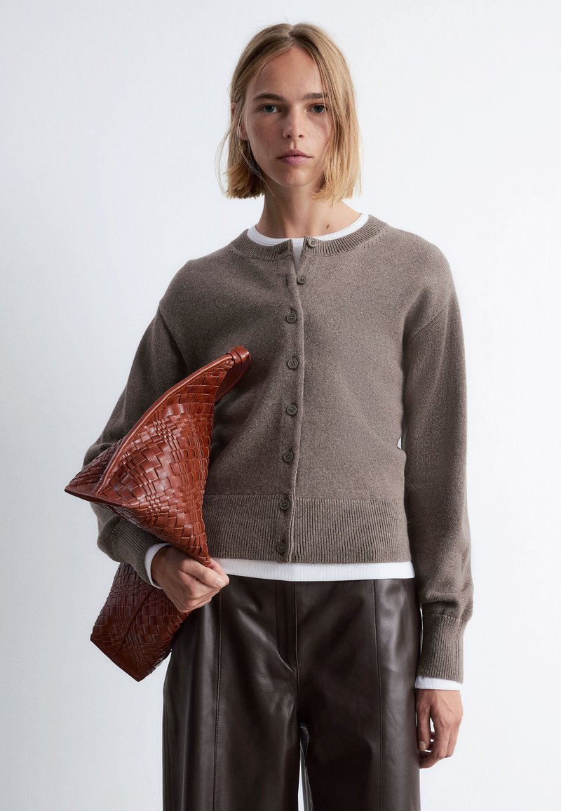 Braune Strickjacke mit rundem Halsausschnitt, fünf Knöpfen und gerippten Bündchen. Gewebte rote Handtasche in der rechten Hand gehalten, Lederhosen sichtbar.