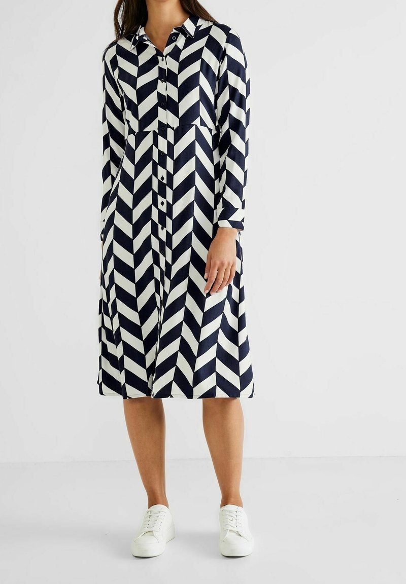 Femme portant une robe longue jusqu'aux genoux, manches longues, boutonnée, avec des rayures chevrons bleu marine et blanc, et des baskets blanches, debout sur un fond uni.