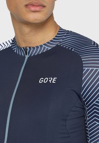 Marin cykeltröja med dragkedja i kragen, GORE-logotyp på bröstet och blå geometrisk mönstring på axlarna. Tillverkad av elastiskt, ventilerande material.