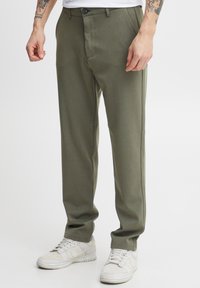 Olive green tailored trousers with a smooth texture, featuring a button closure, side pockets, and a straight leg design, paired with white sneakers.
