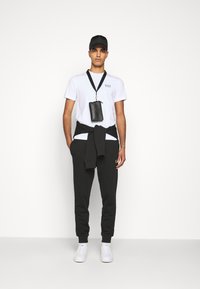 Camiseta blanca con el logo de EA7, pantalones jogger negros y zapatillas blancas. Los accesorios incluyen un bolso cruzado negro y una gorra negra.