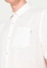 Weißes Leinenhemd mit kurzen Ärmeln und Knopfkragen, ausgestattet mit einer einzelnen Brusttasche und kleinem Palmenmotiv, leichter Stoff.