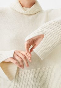 Off-white stickad tröja med hög, ribbad krage, vida ärmar och öppna tumspringor. Texturen är mjuk och mysig med ett horisontellt randmönster.