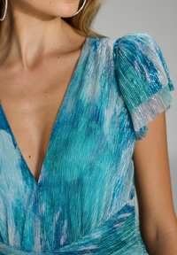 Robe plissée avec un décolleté en V profond, tout en mettant en avant un tissu scintillant teal et bleu. Les manches courtes présentent une finition argentée texturée.