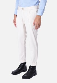 Pantaloni color crema con vita piatta, pieghe e orli risvoltati, abbinati a stivali neri in pelle alla caviglia e una camicia azzurra chiara.