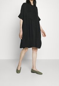 Czarna sukienka z krótkimi rękawami i luźnym krojem, z teksturowanym wzorem; zestawiona z zielonymi płaskimi butami.