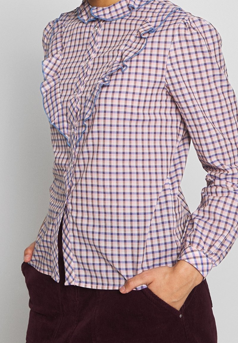 Femme portant un chemisier à carreaux rose, violet et blanc avec des volants et des boutons, associé à un pantalon en velours côtelé violet foncé.