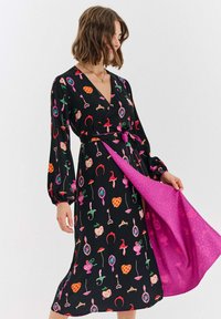 Czarna sukienka kopertowa z kolorowymi wzorami serc, kluczy i koron; ma długie rękawy i różowy akcent w podszewce spódnicy.