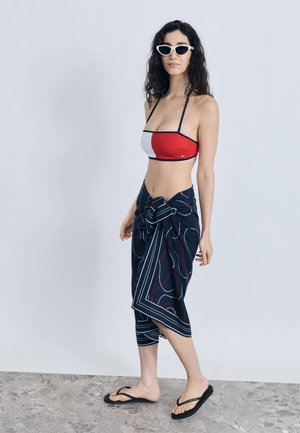 Kobieta w czerwono-białym crop topie, granatowym sarongu z wzorem związanym w talii, czarnych klapkach i białych okularach przeciwsłonecznych w kształcie kociego oka, stojąca na szarej podłodze.