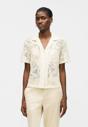 Kvinde iført cremefarvet bluse med knapper, korte ærmer, krave og brystlomme, kombineret med cremefarvede bukser, stående foran en ensfarvet baggrund.