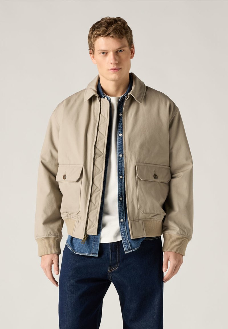 Νεαρός άνδρας που φοράει μπεζ μπουφάν τύπου bomber πάνω από μπλε πουκάμισο από τζιν και λευκό μπλουζάκι, συνδυασμένο με σκουρόχρωμο μπλε τζιν, στέκεται μπροστά σε απλό φόντο.