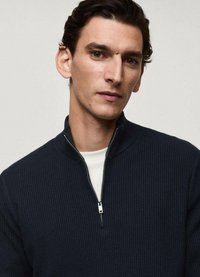 Navy ribbed zip-up sweater featuring a stand collar and silver zipper, worn over a white crew neck shirt. Smooth texture and fitted design.
