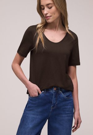 Femme portant un t-shirt marron foncé à manches courtes et col rond, légèrement rentré dans un jean en denim bleu, avec une main dans la poche.