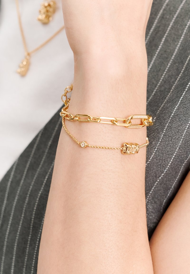 Goldener Gliederarmband mit einer Mischung aus großen Gliedern und zarten Ketten, mit einem kleinen Anhänger und einem Edelsteinakzent. Glatte metallische Textur.