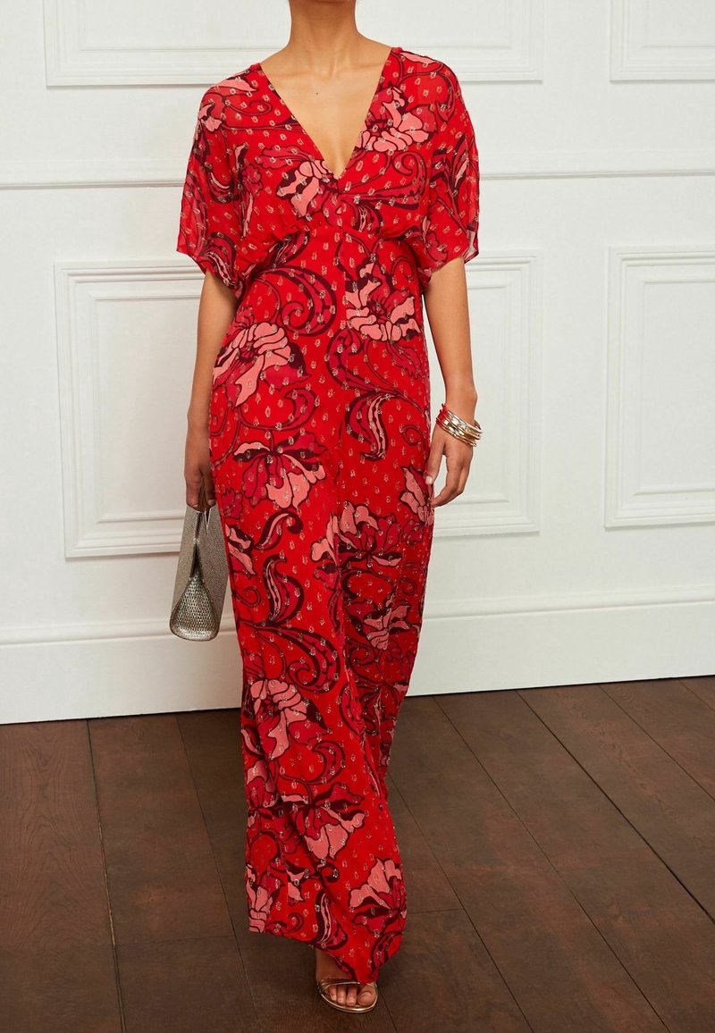 Robe maxi rouge avec des motifs floraux et paisley, décolleté en V profond, manches courtes et coupe ample. Associée à un sac à main métallique et des sandales à lanières.
