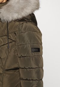 Olivgrüner, gesteppten Winterjacke mit einer Kapuze mit Pelzbesatz und Kordelzug, die ein schwarzes DKNY-Logo-Patch am Ärmel aufweist.