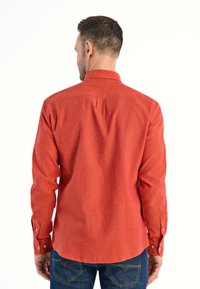 Czerwona, długosleeve koszula zapinana na guziki o teksturowanym wykończeniu i plisie z tyłu. Ma zaokrąglony dół i guziki przy mankietach, zestawiona z niebieskimi dżinsami.